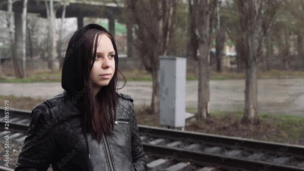 A young woman stands on the platform and waiting for the train. Female in a leather jacket with a hood, looking away on a rainy day. A young woman stands on the platform and waiting for the train. Female in a leather jacket with a hood, looking away on a rainy day.
