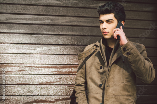 Portrait of a model young male Turkish Mediterranean race brunet in coat uses hand phone technology businessman on the street in sunny weather against the background of a wooden wall photo