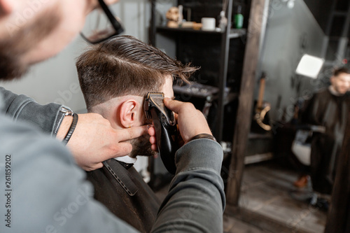 Master cuts hair and beard of men in the barbershop, hairdresser makes hairstyle for a young man. Barber work with clipper machine in barbershop.