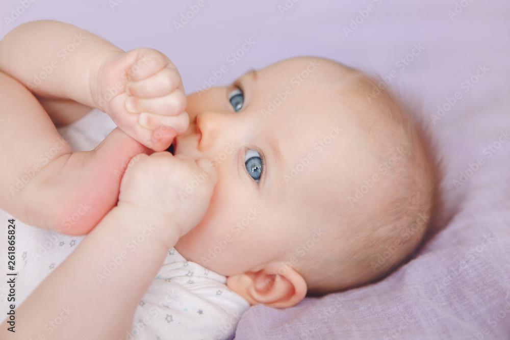 Closeup portrait of cute adorable white Caucasian infant child girl boy with blue eyes lying on bed licking sucking fingers fist. Baby teething. Happy childhood lifestyle and newborn development. Closeup portrait of cute adorable white Caucasian infant child girl boy with blue eyes lying on bed licking sucking fingers fist. Baby teething. Happy childhood lifestyle and newborn development.
