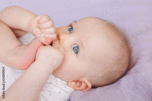 Closeup portrait of cute adorable white Caucasian infant child girl boy with blue eyes lying on bed licking sucking fingers fist. Baby teething. Happy childhood lifestyle and newborn development. © anoushkatoronto