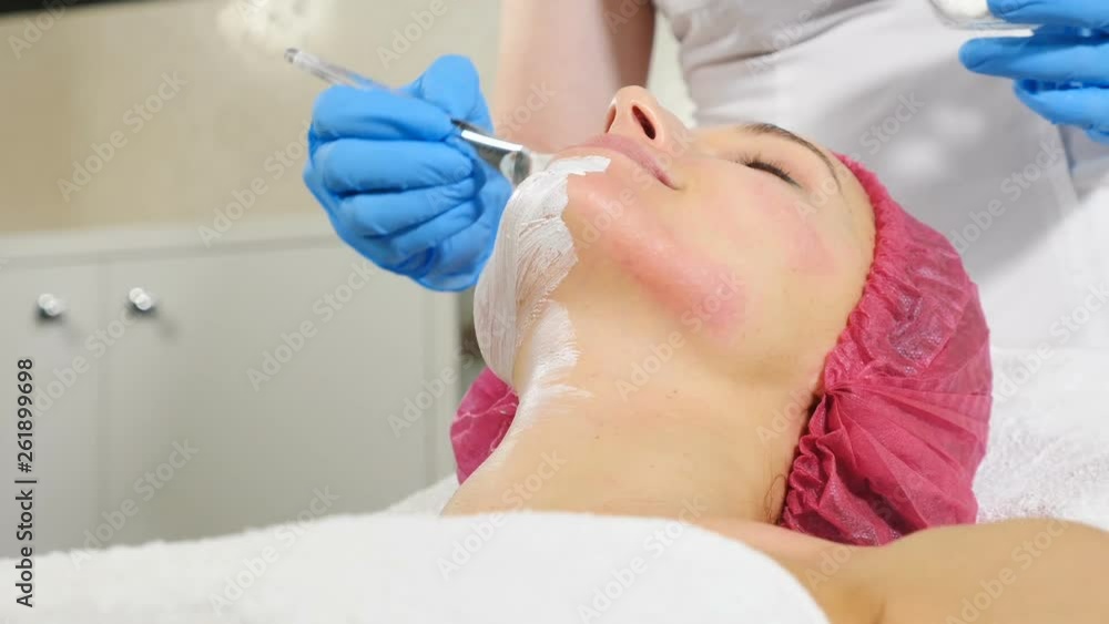 Attractive female client with facial mask at beauty salon.Applying facial mask to female face with a brush at dermatology clinic. Spa therapy procedure for young woman.Beautician applying facial mask Attractive female client with facial mask at beauty salon.Applying facial mask to female face with a brush at dermatology clinic. Spa therapy procedure for young woman.Beautician applying facial mask