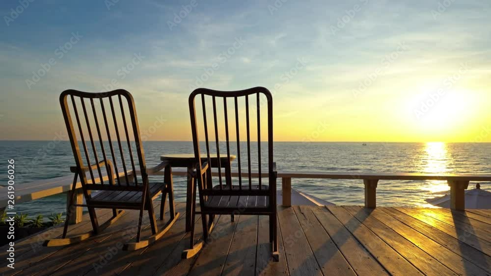 Beautiful and relaxing ocean sunset. Two empty rocking chairs on a wooden deck and golden light of the setting sun reflected on a calm sea. Peaceful evening scene with no people. Beautiful and relaxing ocean sunset. Two empty rocking chairs on a wooden deck and golden light of the setting sun reflected on a calm sea. Peaceful evening scene with no people.