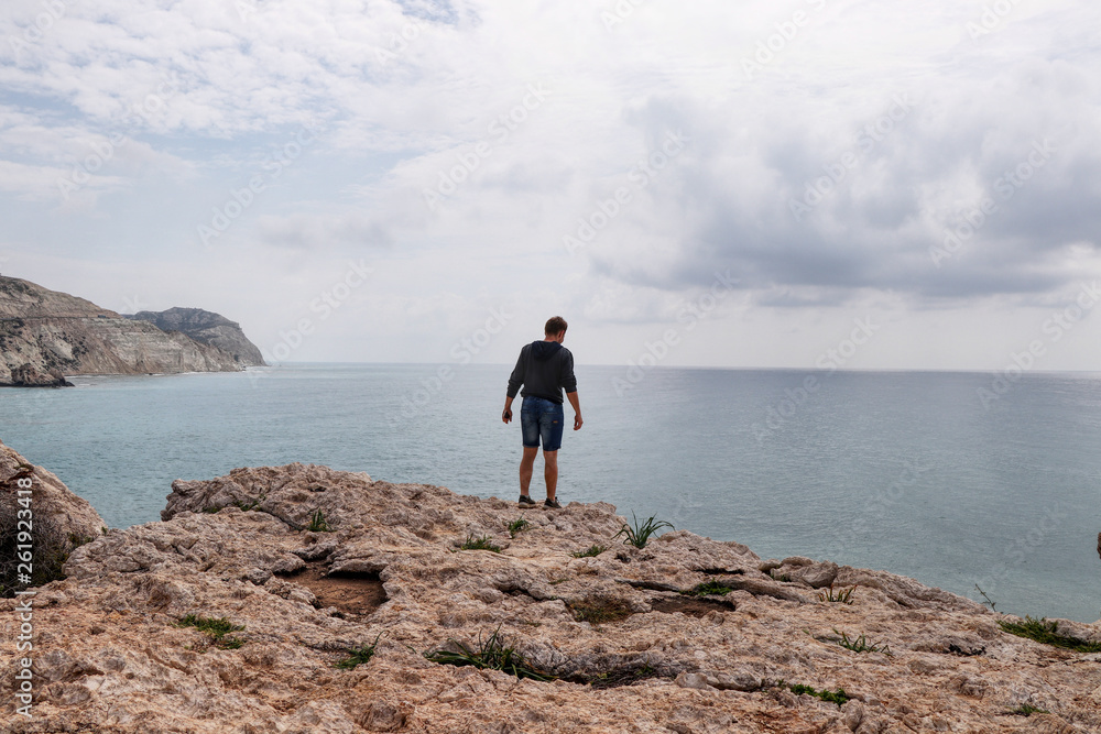 Teenager on the edge of rock Of Greek and watchs waves down. Man is on Aphrodite's rock on Petra de romiou beach. Adolescent climbs on big hill and enjoy scenery and panorama Teenager on the edge of rock Of Greek and watchs waves down. Man is on Aphrodite's rock on Petra de romiou beach. Adolescent climbs on big hill and enjoy scenery and panorama