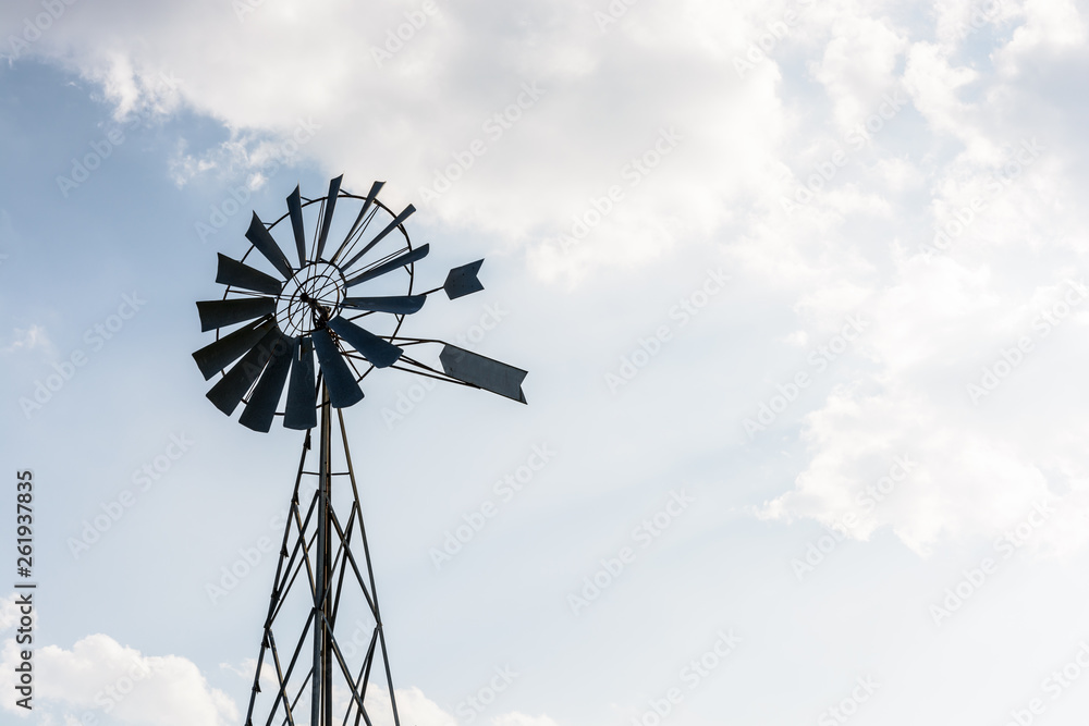 Low angle view of an old-fashioned, multi-bladed, metal wind powered water pump atop a lattice tower in backlight against a pale blue sky with white clouds. Low angle view of an old-fashioned, multi-bladed, metal wind powered water pump atop a lattice tower in backlight against a pale blue sky with white clouds.