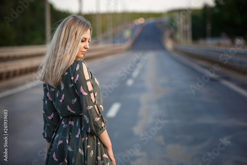 Young blonde girl on the road in the evening soft light of the sun. A girl in a green dress with long hair, the dress is blown by the wind.