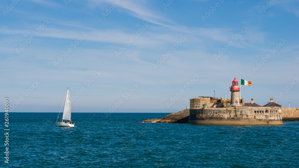 Sail boat leaving the harbor. The lighthouse of Dun Laoghaire East Pier on a sunny summer day. Seascape in Dublin, Ireland.