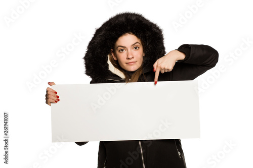 Young model in winter jacket with hood shows an empty advertising board on white background
