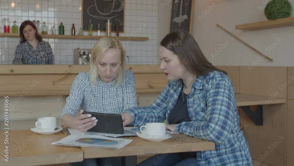 Serious females entrepreneurs working remotely brainstorming during start-up of business project. Confident women freelance using tablet and charts communicating at meeting in coffee shop. Serious females entrepreneurs working remotely brainstorming during start-up of business project. Confident women freelance using tablet and charts communicating at meeting in coffee shop.