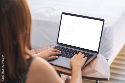 Mockup image of a woman using and typing on laptop with blank white desktop screen keyboard on wooden table next to the bed