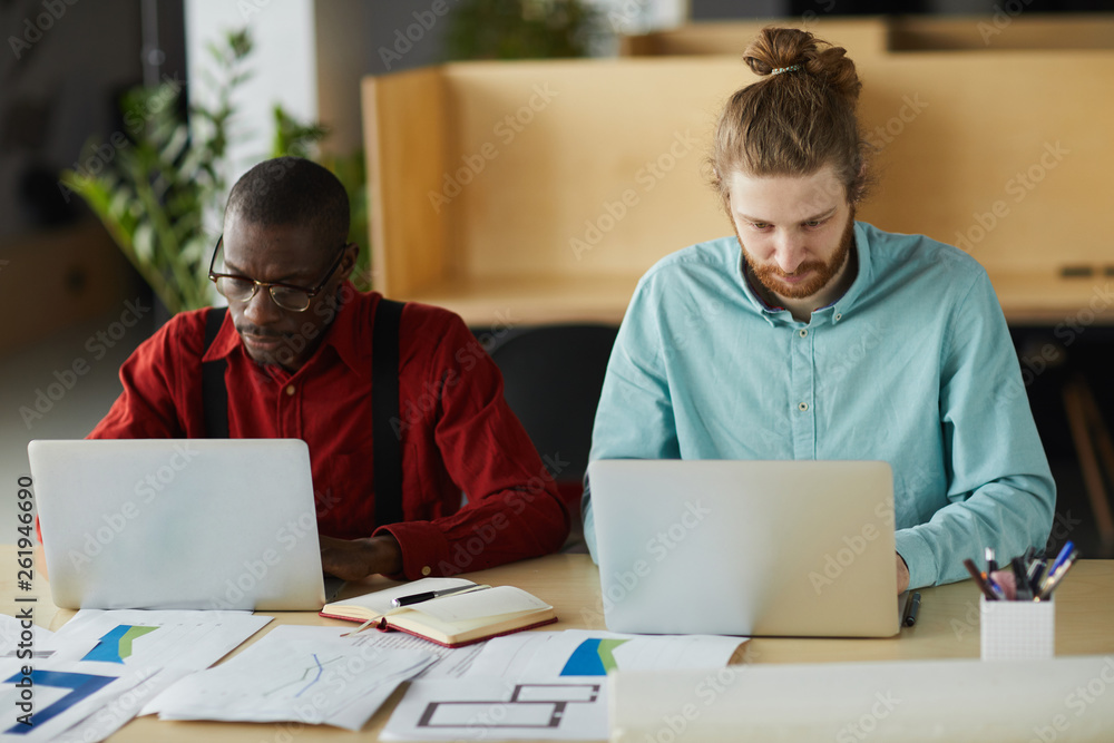 Obraz premium Portrait of two contemporary business using laptops sitting at desk and while working in office