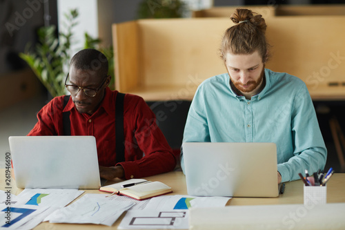 Portrait of two contemporary business using laptops sitting at desk and while working in office