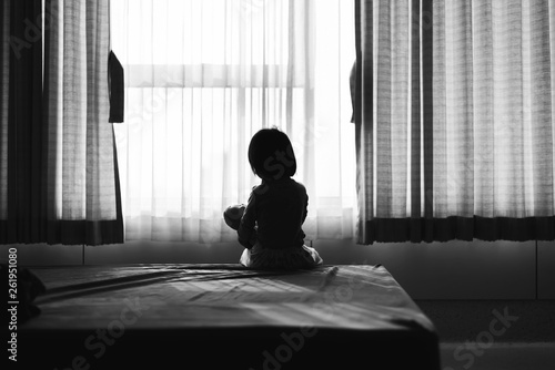 Little baby child sitting on bed playing with the doll in the dark room with light throught curtain
