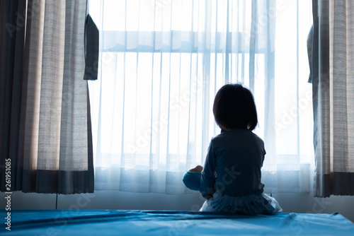 Little baby child sitting on bed playing with the doll in the dark room with light throught curtain photo