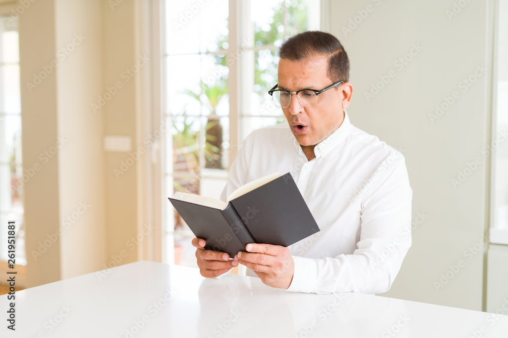 Middle age man reading a book at home wearing glasses scared in shock with a surprise face, afraid and excited with fear expression Middle age man reading a book at home wearing glasses scared in shock with a surprise face, afraid and excited with fear expression