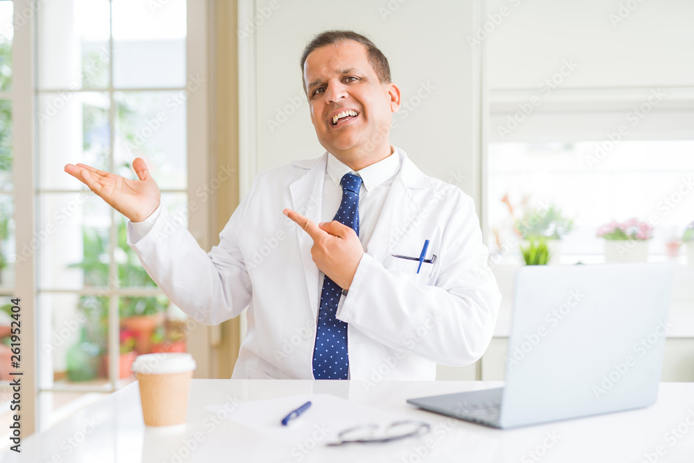 Middle age doctor man wearing white medical coat working with laptop at the clinic amazed and smiling to the camera while presenting with hand and pointing with finger. Middle age doctor man wearing white medical coat working with laptop at the clinic amazed and smiling to the camera while presenting with hand and pointing with finger.