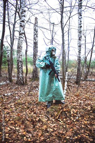 Stalker, a man in a gas mask and special chemical protection, green cloak.  With a gun in his hand, walking in the forest © Vera