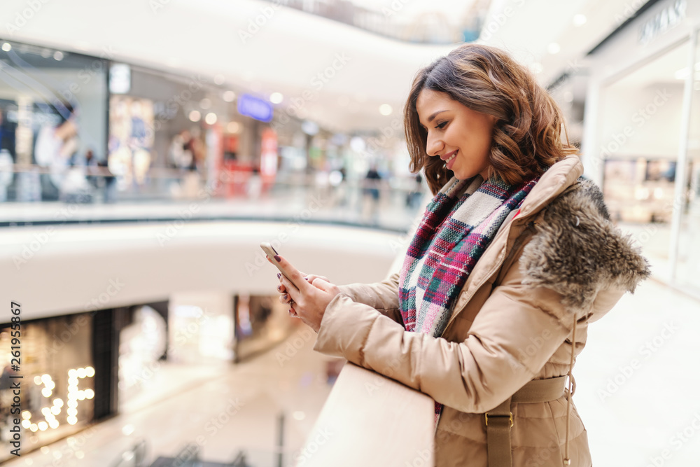 Beautiful smiling brunette in jacket and with scarf leaning on the fence and using smart phone for writing message. Shopping mall interior.