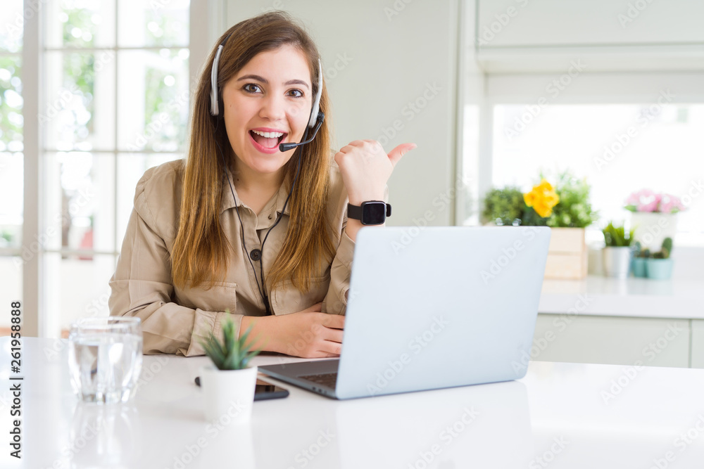 Beautiful young operator woman working with laptop and wearing headseat smiling with happy face looking and pointing to the side with thumb up. Beautiful young operator woman working with laptop and wearing headseat smiling with happy face looking and pointing to the side with thumb up.