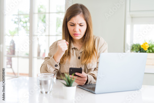 Beautiful young woman using smartphone and computer annoyed and frustrated shouting with anger, crazy and yelling with raised hand, anger concept