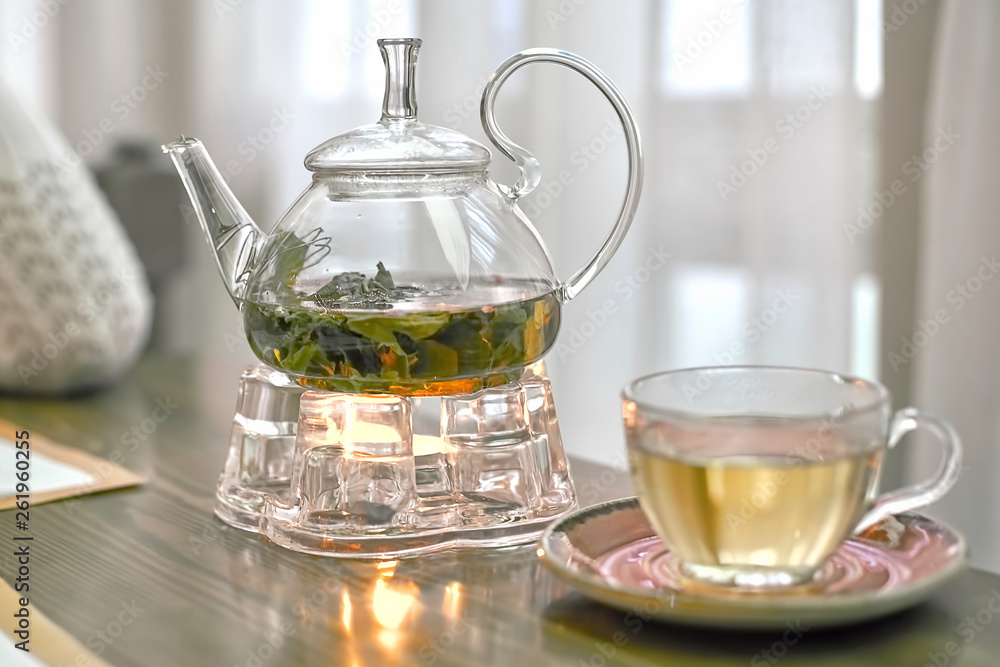 Transparent teapot with green tea on a stand with a candle, close-up Transparent teapot with green tea on a stand with a candle, close-up