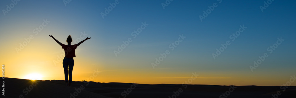 Silhouette of a woman standing in the sunset with the arms up, panoramic background with copy space