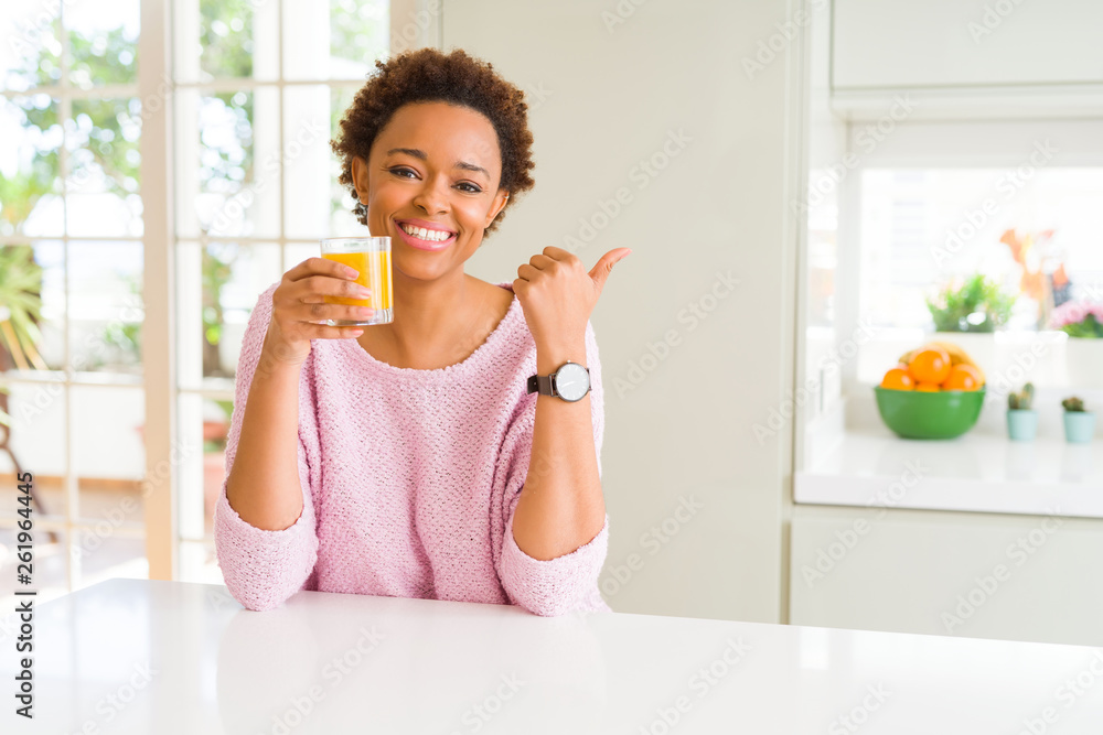 Young african american woman driking orange juice at home pointing and showing with thumb up to the side with happy face smiling Young african american woman driking orange juice at home pointing and showing with thumb up to the side with happy face smiling