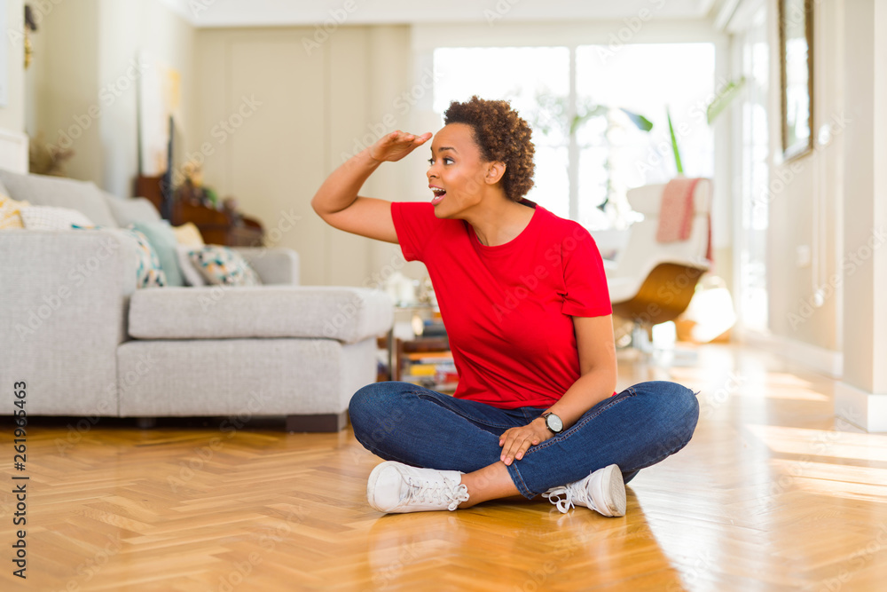 Young beautiful african american woman sitting on the floor at home very happy and smiling looking far away with hand over head. Searching concept.