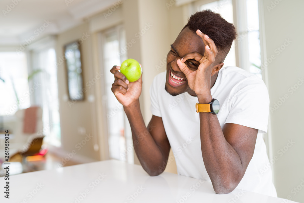 Young african american man eating fresh green apple with happy face smiling doing ok sign with hand on eye looking through fingers Young african american man eating fresh green apple with happy face smiling doing ok sign with hand on eye looking through fingers