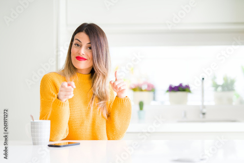 Young beautiful woman drinking a cup of coffee at home pointing fingers to camera with happy and funny face. Good energy and vibes.
