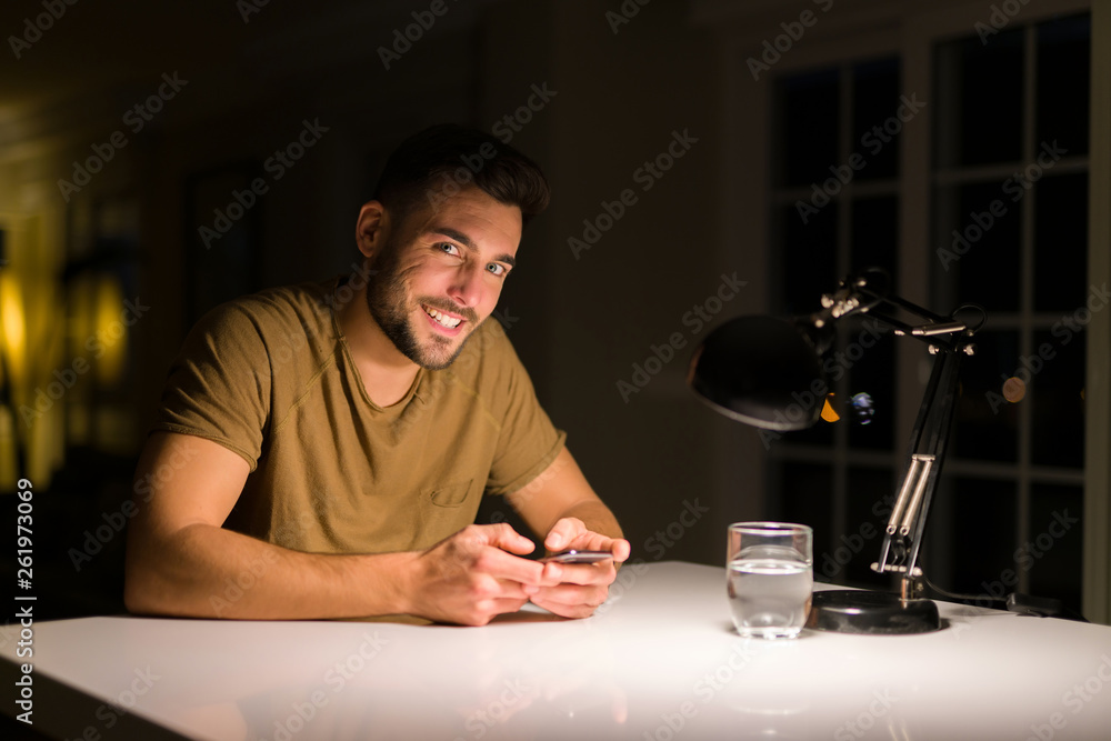 Young handsome man studying at home , using smartphone, smiling at the camera Young handsome man studying at home , using smartphone, smiling at the camera