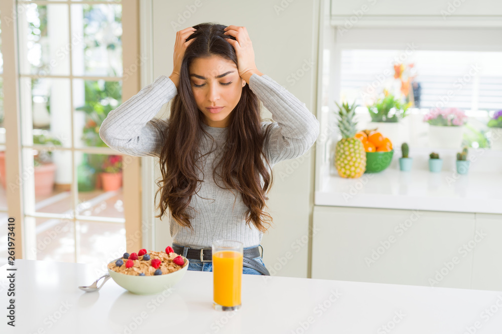 Young woman eating healthy breakfast in the morning suffering from headache desperate and stressed because pain and migraine. Hands on head. Young woman eating healthy breakfast in the morning suffering from headache desperate and stressed because pain and migraine. Hands on head.