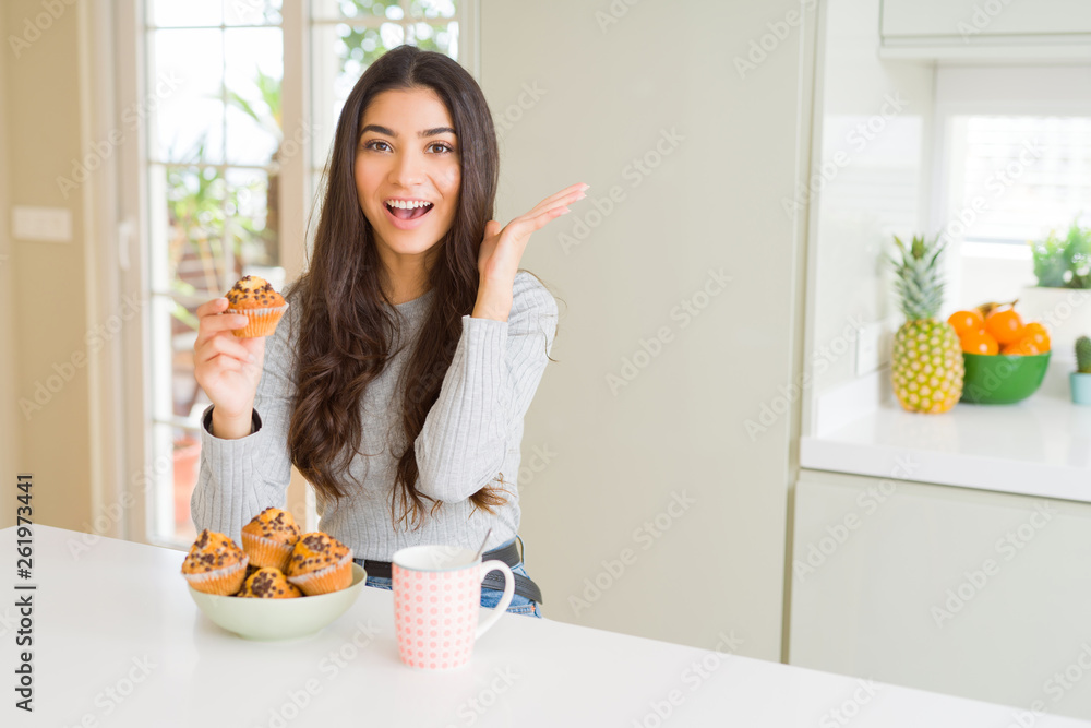 Young woman eating chocolate chips muffins and drinking coffee very happy and excited, winner expression celebrating victory screaming with big smile and raised hands Young woman eating chocolate chips muffins and drinking coffee very happy and excited, winner expression celebrating victory screaming with big smile and raised hands