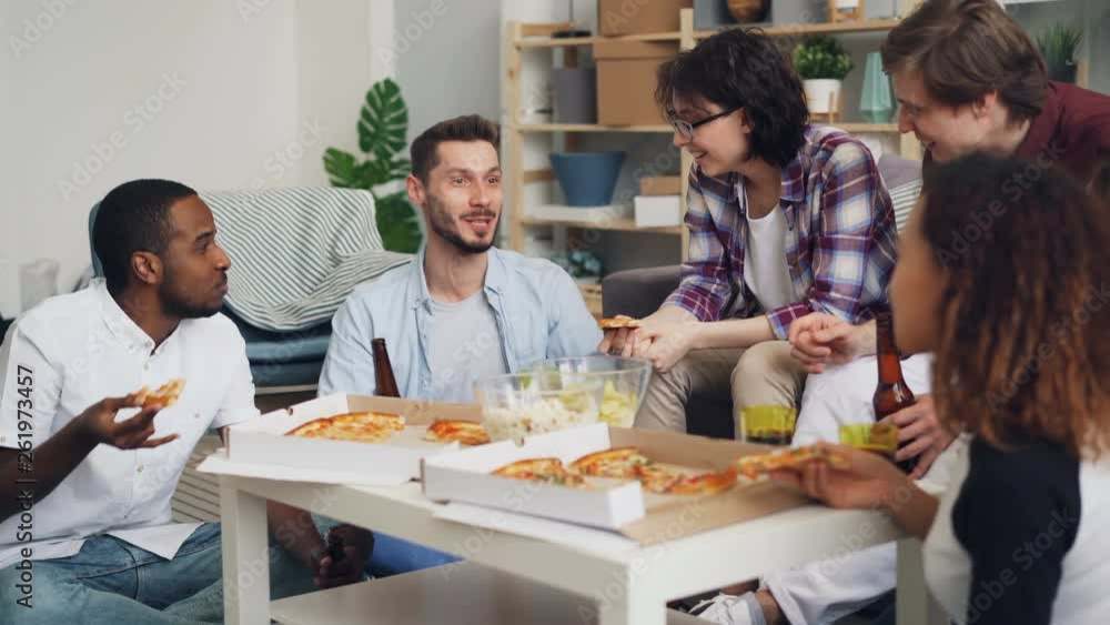 Girls and guys are toasting and clinking bottle and glasses eating pizza chatting and laughing at home party together. Celebration and indoor activities concept. Girls and guys are toasting and clinking bottle and glasses eating pizza chatting and laughing at home party together. Celebration and indoor activities concept.