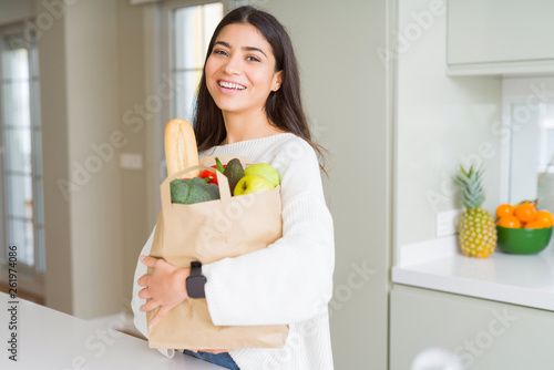 Beautiful young woman smiling holding a paper bag full of groceries at the kitchen