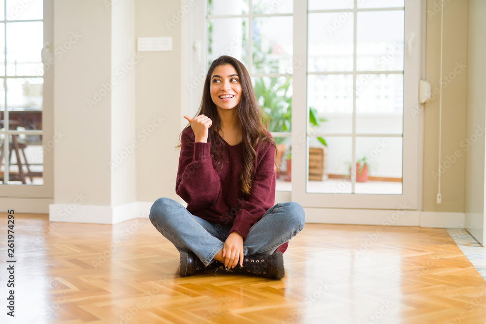Young beautiful woman sitting on the floor at home smiling with happy face looking and pointing to the side with thumb up.