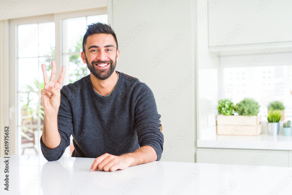 Handsome hispanic man wearing casual sweater at home showing and pointing up with fingers number three while smiling confident and happy.