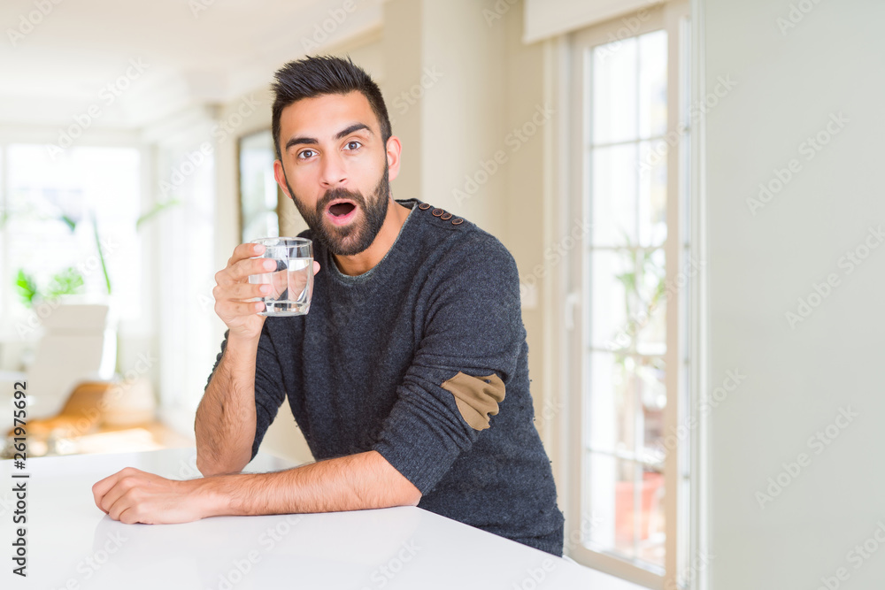 Handsome hispanic man drinking a fresh glass of water scared in shock with a surprise face, afraid and excited with fear expression