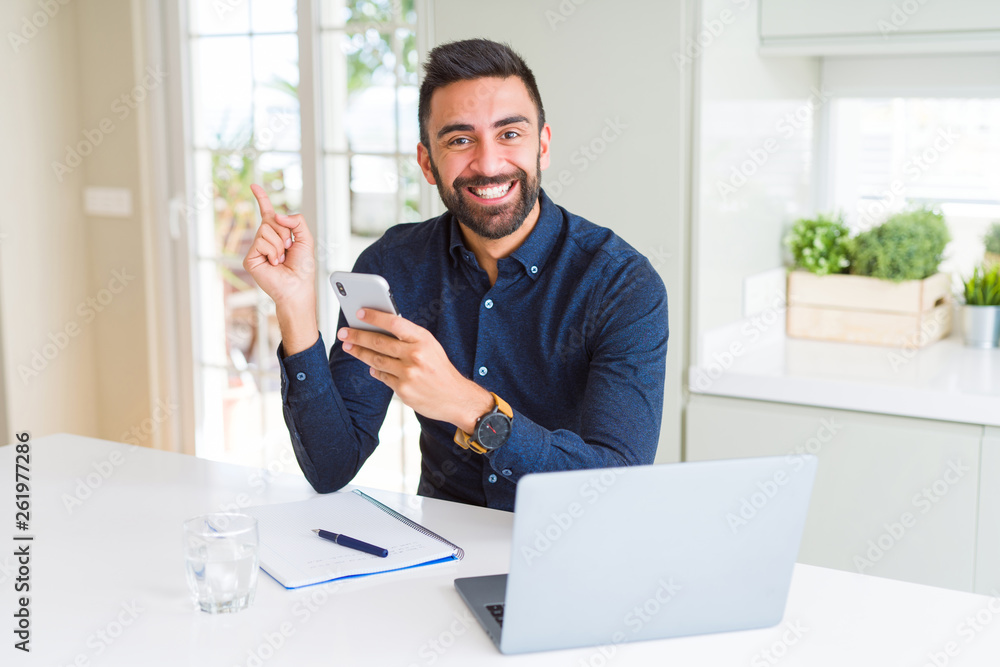 Handsome hispanic business man using smartphone and laptop at the office very happy pointing with hand and finger to the side