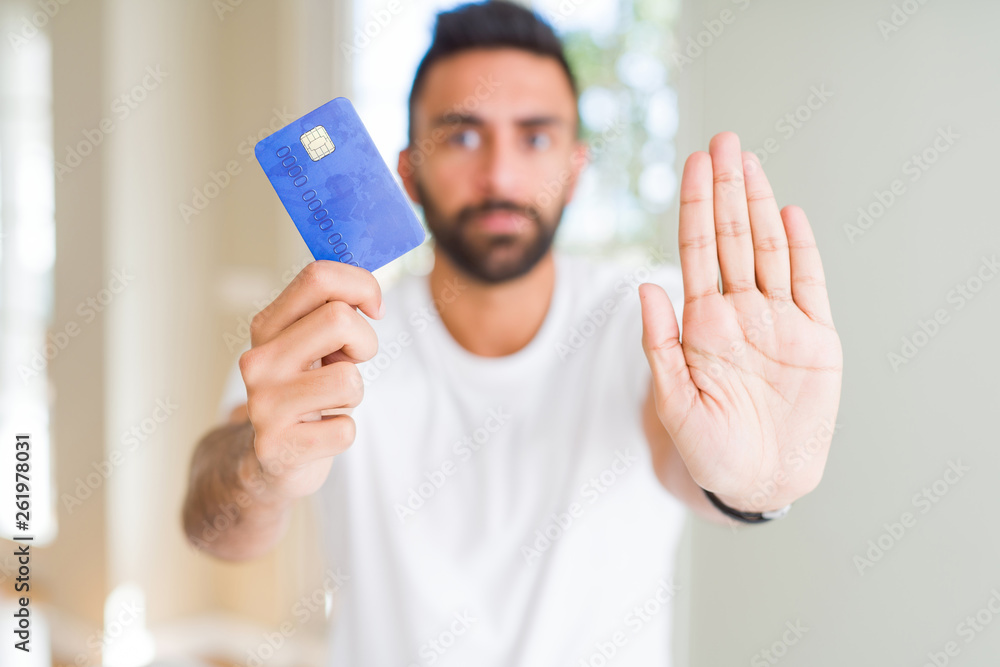 Handsome hispanic man holding credit card with open hand doing stop sign with serious and confident expression, defense gesture Handsome hispanic man holding credit card with open hand doing stop sign with serious and confident expression, defense gesture