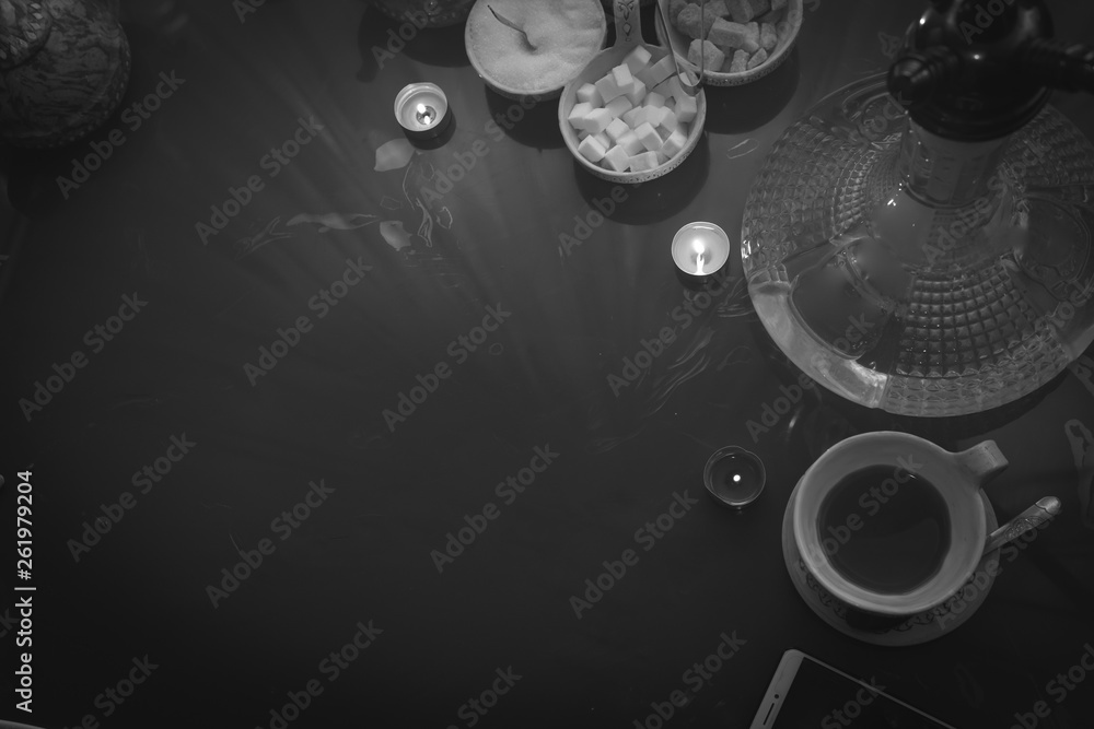 Obraz premium Hookah on glass table top view. cups candle and smartphone eastern tea ceremony. Selective focus, sugar cubes with different flavors. Copy space, black and white