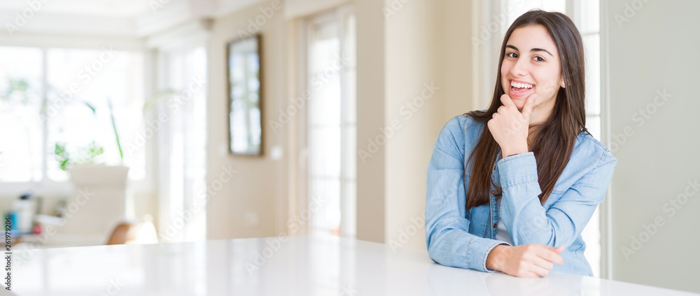 Wide angle picture of beautiful young woman sitting on white table at home looking confident at the camera with smile with crossed arms and hand raised on chin. Thinking positive. Wide angle picture of beautiful young woman sitting on white table at home looking confident at the camera with smile with crossed arms and hand raised on chin. Thinking positive.