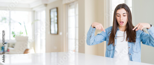 Wide angle picture of beautiful young woman sitting on white table at home Pointing down with fingers showing advertisement, surprised face and open mouth