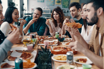 Feeling hungry. Group of young people in casual wear eating pizza and smiling while having a dinner party indoors