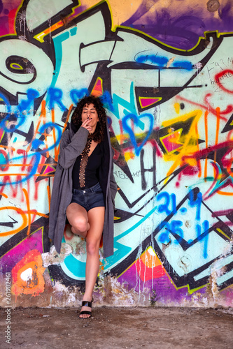 Girl with black curly hair and with a cigarette in her mouth leaning against the wall with graffiti