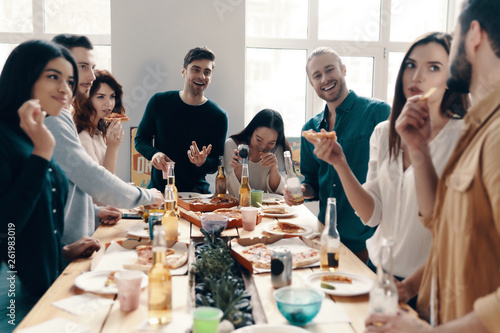 Pizza party. Group of young people in casual wear eating pizza and smiling while having a dinner party indoors