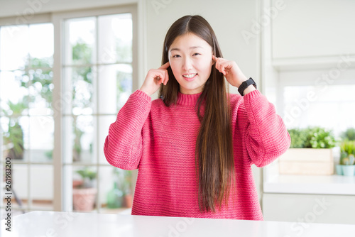 Beautiful Asian woman wearing pink sweater on white table covering ears with fingers with annoyed expression for the noise of loud music. Deaf concept.