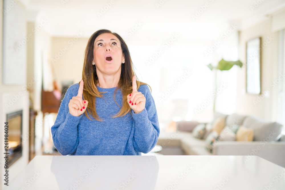 Beautiful middle age woman at home amazed and surprised looking up and pointing with fingers and raised arms. Beautiful middle age woman at home amazed and surprised looking up and pointing with fingers and raised arms.