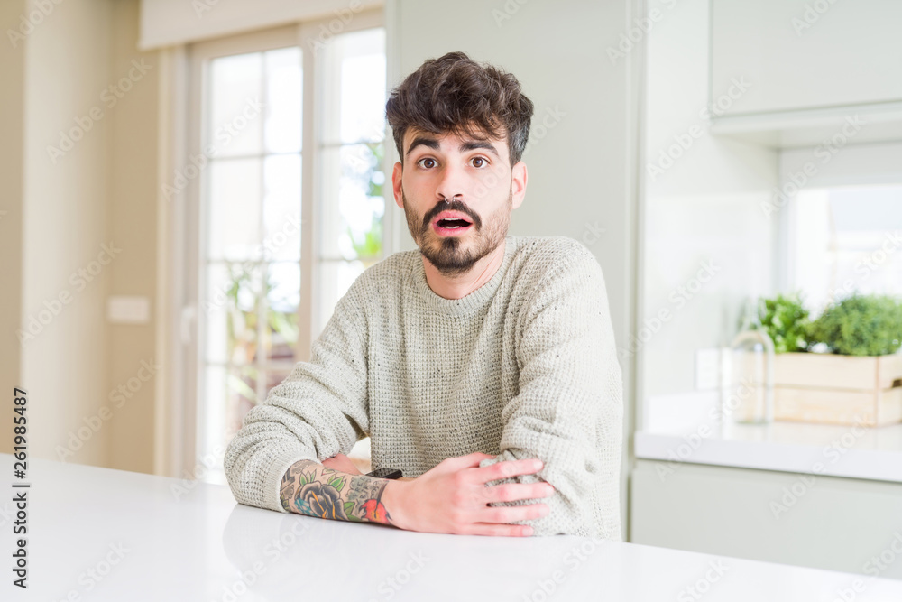 Young man wearing casual sweater sitting on white table afraid and shocked with surprise expression, fear and excited face.