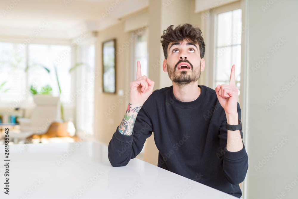 Young man wearing casual sweatshirt sitting on white table amazed and surprised looking up and pointing with fingers and raised arms. Young man wearing casual sweatshirt sitting on white table amazed and surprised looking up and pointing with fingers and raised arms.